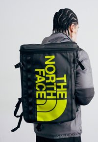 Sac à dos noir avec un grand logo jaune "THE NORTH FACE", design rectangulaire, fermeture par zip en haut et bretelles réglables.