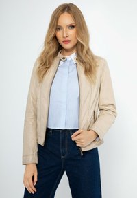 Beige kožená bunda s přiléhavým střihem, se zipem a stojáčkem. Nositá přes světle modrou pruhovanou blůzu a tmavé džíny s vysokým pasem.