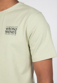 Světle zelené bavlněné tričko se černě potisknutým logem "WRONG FRIENDS" na levé horní straně, s klasickým kulatým výstřihem a krátkými rukávy.