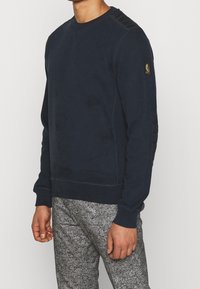 Man som bär en marinblå sweatshirt med lappdetaljer på axeln och ärmen, ihop med texturerade grå byxor, står mot en enkel bakgrund.