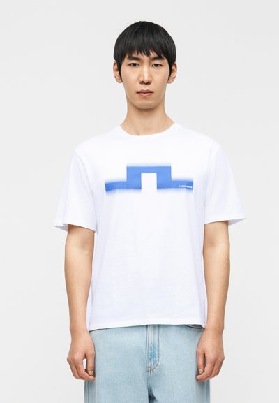 Άνδρας που φοράει λευκό T-shirt με μπλε γεωμετρικό σχέδιο και ανοιχτόχρωμο μπλε τζιν, στέκεται μπροστά σε μία απλή φωτεινή επιφάνεια.