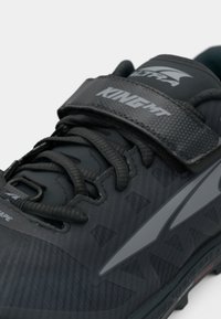 Black athletic shoe with textured upper, adjustable strap, and grey accents. Features enclosed lacing system and rugged sole pattern.