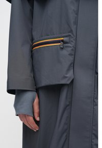 Sivé vonkajšie oblečenie so zipsovým vreckom s farebným prúžkom, zúženými rukávmi a voľným strihom. Materiál pôsobí hladko a ľahko.