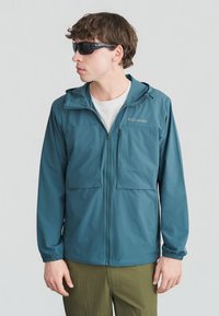 Νεαρός άνδρας που φοράει τζάκετ Columbia σε χρώμα τιρκουάζ με φερμουάρ, λευκό πουκάμισο, ελαιώδη παντελόνια και μαύρα γυαλιά ηλίου τύπου wraparound, κοιτάζει αριστερά με φόντο απλό.