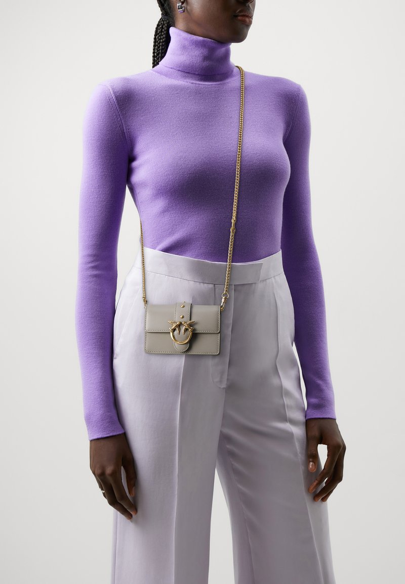 Lilla tettsittende turtleneck genser i kombinasjon med lyse grå bukser. Tilbehøret inkluderer en liten grå crossover-veske med gullkjede og dekorativt hardware.