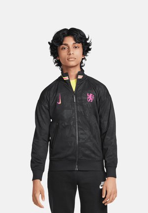 Veste zippée noire en tissu léger, dotée d'un motif subtil, d'un logo Nike rose et d'un blason. Col en rib avec des accents colorés.
