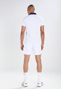 Hvit polo skjorte med vertikale marineblå striper, marineblå krage, kombinert med hvite shortser og knelange hvite sokker, båret av en person sett bakfra.