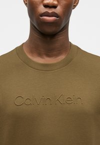 Κοντινό πλάνο ενός ατόμου που φορά ένα ελιάς πράσινο πουκάμισο με ανάγλυφο κείμενο "Calvin Klein" στο στήθος, εμφανίζοντας τον λαιμό και το κάτω μέρος του προσώπου.