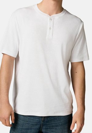 T-shirt henley à manches courtes blanc en coton côtelé, avec un col rond et une patte de boutonnage à deux boutons. Porté avec un jean bleu.