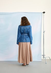 Femeie cu păr creț poartă o jachetă din denim legată, o fustă lungă bej și pantofi cu dungi, stând cu fața spre un fundal albastru deschis.