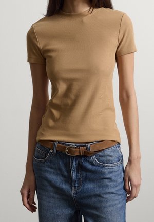 Kvinde iført en tætsiddende beige ribbet top med korte ærmer, stoppet ned i højttaljede blå jeans med et brunt læderbælte.