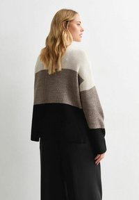 Femme avec de longs cheveux blonds portant un pull en maille en blocs de couleurs blanc, taupe et noir, se tenant de côté contre un mur clair et uni.