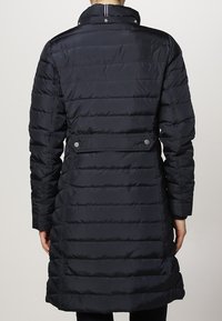 Marineblauwe donsjas met een quilted ontwerp, een ceintuur in de taille, hoge kraag en drukknopen. Soepel, glanzend materiaal; reikt tot halverwege de bovenbenen.