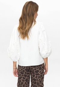 Vrouw met lang bruin haar, gekleed in een witte blouse met opstaande, gerimpelde mouwen en een broek met een bruin patroon, gezien vanuit de achterkant.