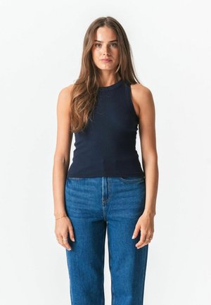 Fiatal nő hosszú barna hajjal, ujjatlan tengerészkék felsőt és kék, magas derekú farmert visel, sima fehér háttér előtt állva.