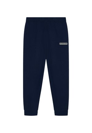 Joven de pie con sudadera negra y pantalones con rayas blancas y zapatillas blancas de caña alta sobre fondo liso.
