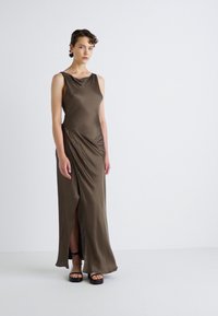 Long, olive green dress made of smooth fabric with a draped design and side slit. Model wears black chunky sandals. Simple, elegant silhouette.