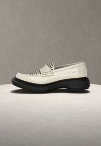 Λευκό μοκασίνι από δέρμα με μαύρη χοντρή σόλα και διάτρητο επάνω μέρος, εκτεθειμένο σε ουδέτερο φόντο πάνω σε στρογγυλή επιφάνεια.