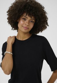 Tricou negru tricotat cu mâneci scurte, guler rotund și o textură netedă. Un brățară argintie pe încheietura mâinii adaugă un accent metalic.