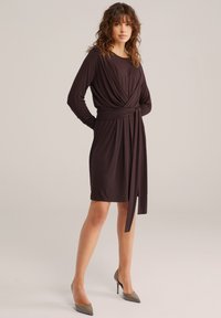 Vrouw staat met handen achter de rug, draagt een donkerkleurige wikkeljurk met lange mouwen tot op de knie en puntige hoge hakken, tegen een effen achtergrond.