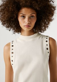Göndör hajú nő, aki ujjatlan krém színű felsőt visel, fémlyukakkal a vállvarrás mellett, és közvetlenül előre néz.