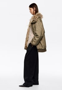 Chaqueta bomber verde oliva con cuello de piel sintética, combinada con un suéter ligero y pantalones anchos negros. Presenta un bolsillo lateral en la chaqueta.