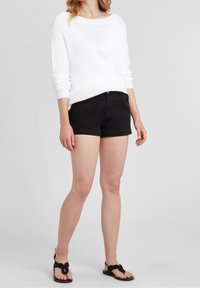 Biały sweter robiony na drutach z teksturowanym wzorem, zestawiony z czarnymi szortami i czarnymi sandałami z detalem w postaci węzła. Prosty i casualowy strój.