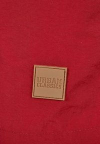 Piros szövet, amelyen egy négyzet alakú, bézs címke van belevarrva, rajta a "URBAN CLASSICS" felirattal, domborítva.