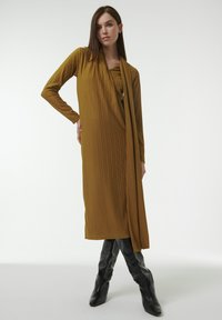 Vestido largo de canalé en color mostaza con escote drapeado y mangas largas. Combinado con botas de cuero negro hasta la rodilla.