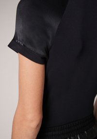 Top negro con un tejido texturizado y mangas satinadas. Diseño estructurado con una silueta ajustada. Detalle de puños enrollados.
