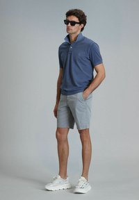 Blaues Polohemd mit umgeschlagenem Kragen, kombiniert mit hellgrauen Shorts. Dazu weiße Sneaker und schwarze Sonnenbrille.