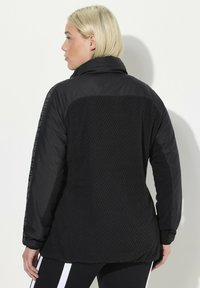 Veste noire avec un dos texturé présentant un motif géométrique, des manches lisses contrastantes et un col haut. Détail du texte logo sur le côté.