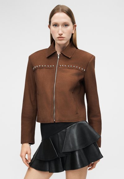 Γυναίκα που φοράει καφέ ζακέτα με φερμουάρ και μεταλλικά τρουκς στο στήθος, και μαύρη δερμάτινη φούστα με βολάν, στέκεται μπροστά σε απλό φόντο.