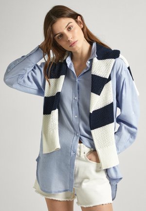 Camicia blu a maniche lunghe con maniche arrotolate abbinata a una sciarpa di maglia a righe in blu navy e bianco. Pantaloni corti bianchi completano l'outfit.