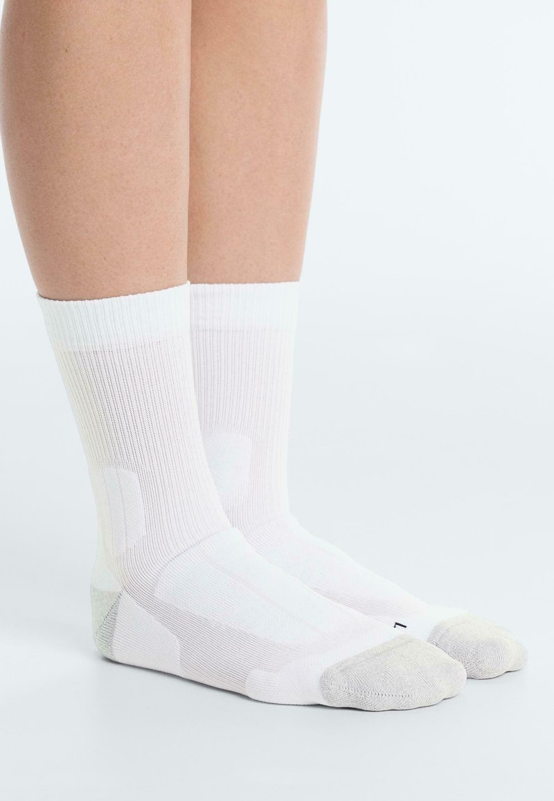 Paire de jambes nues portant des chaussettes blanches montantes avec des zones renforcées grises aux orteils et aux talons, debout sur une surface blanche.