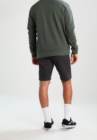 Felpa girocollo verde scuro, pantaloni corti neri, calzini bianchi con logo e scarpe da ginnastica nere. Outfit informale, vista da dietro. Texture del tessuto liscia.
