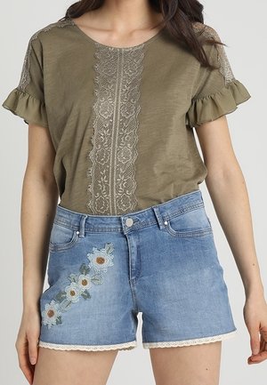 Woman wearing olive green short-sleeve top with lace details and light blue denim shorts featuring white floral embroidery and lace trim.