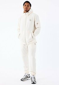 Conjunto de sudadera y pantalones color crema, con bolsillos con cremallera, logotipos y tela texturizada. Combinado con zapatillas deportivas negras y plateadas.