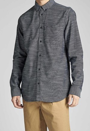 Mann trägt ein dunkelgraues Hemd mit langen Ärmeln und Brusttasche sowie beige Hosen, steht vor einem schlichten Hintergrund.