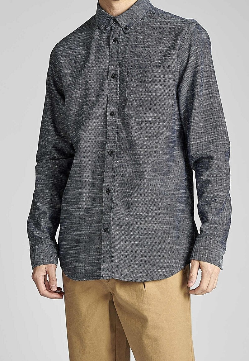 Homme portant une chemise à manches longues gris foncé avec une poche poitrine et un pantalon beige, debout devant un fond uni.