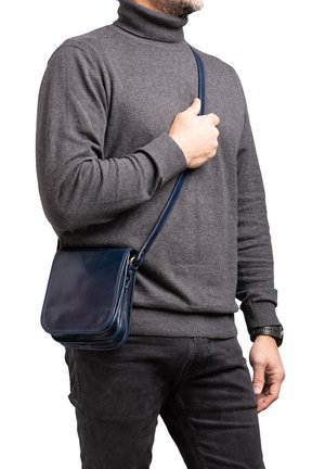 Borsa a tracolla in pelle blu navy con finitura liscia, forma quadrata e tracolla regolabile, tenuta da una persona che indossa un maglione grigio a collo alto.