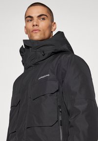 Svart vattentät jacka med hög krage, flera fickor och dragkedja. Har en matt yta och diskret märkning.