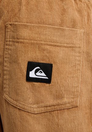 Brązowa kieszeń z sztruksu z naszywką z logo Quiksilver w czarno-białych kolorach, przyszytą na środku kieszeni.