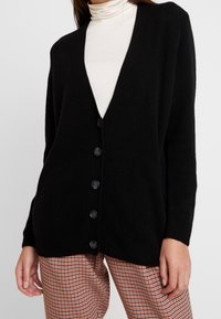 Svart cardigan med V-ringning, sju stora knappar fram och ribbade ärmar; bärs över en krämfärgad turtleneck.