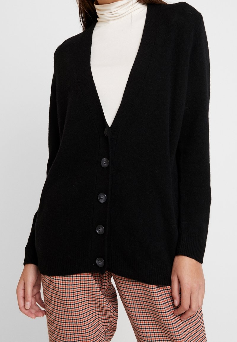 Svart cardigan med V-ringning, sju stora knappar fram och ribbade ärmar; bärs över en krämfärgad turtleneck.