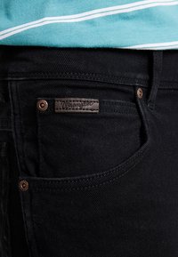 Gros plan d'une poche de jean en denim noir avec un patch en cuir de la marque Wrangler et des rivets en cuivre, porté avec un tissu rayé bleu sarcelle et blanc au-dessus.