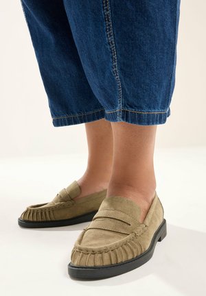 Personne portant des mocassins en daim beige et un pantalon en denim bleu raccourci, debout sur un sol de couleur claire.