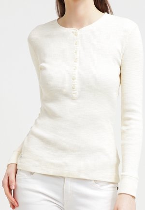 Femme portant un t-shirt Henley ajusté à manches longues couleur crème avec des boutons et un pantalon blanc, main dans la poche, fond neutre.
