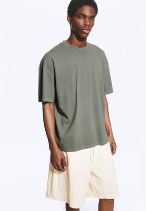 Jeune homme portant un t-shirt oversize vert olive et un short beige ample jusqu'aux genoux, debout devant un fond blanc uni.