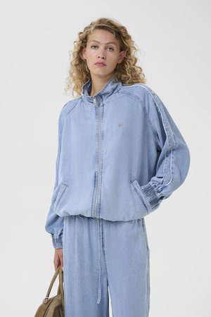 Γυναίκα με σγουρά μαλλιά, φοράει ανοιχτό μπλε τζιν μπουφάν και παντελόνι, κρατώντας καφέ τσάντα, κοιτάζοντας την κάμερα μπροστά σε απλό γκρι φόντο.